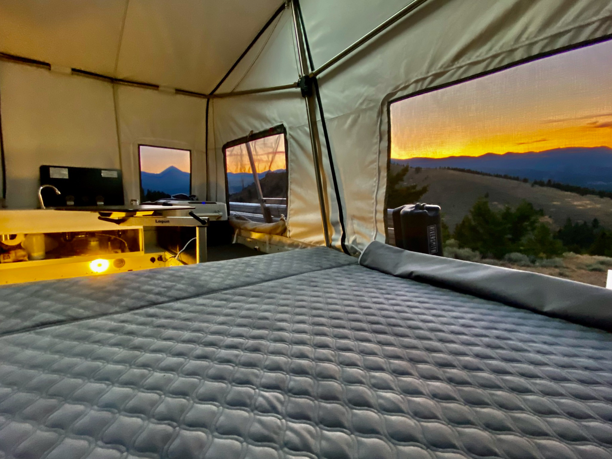 Interior of a tent at sunset, featuring a neatly made bed with a quilted cover. Two large windows reveal a vibrant sunset over rolling hills and trees. In the background, a camping stove and cooking supplies are visible on a counter, making it an ideal setup for overland campers seeking comfort.