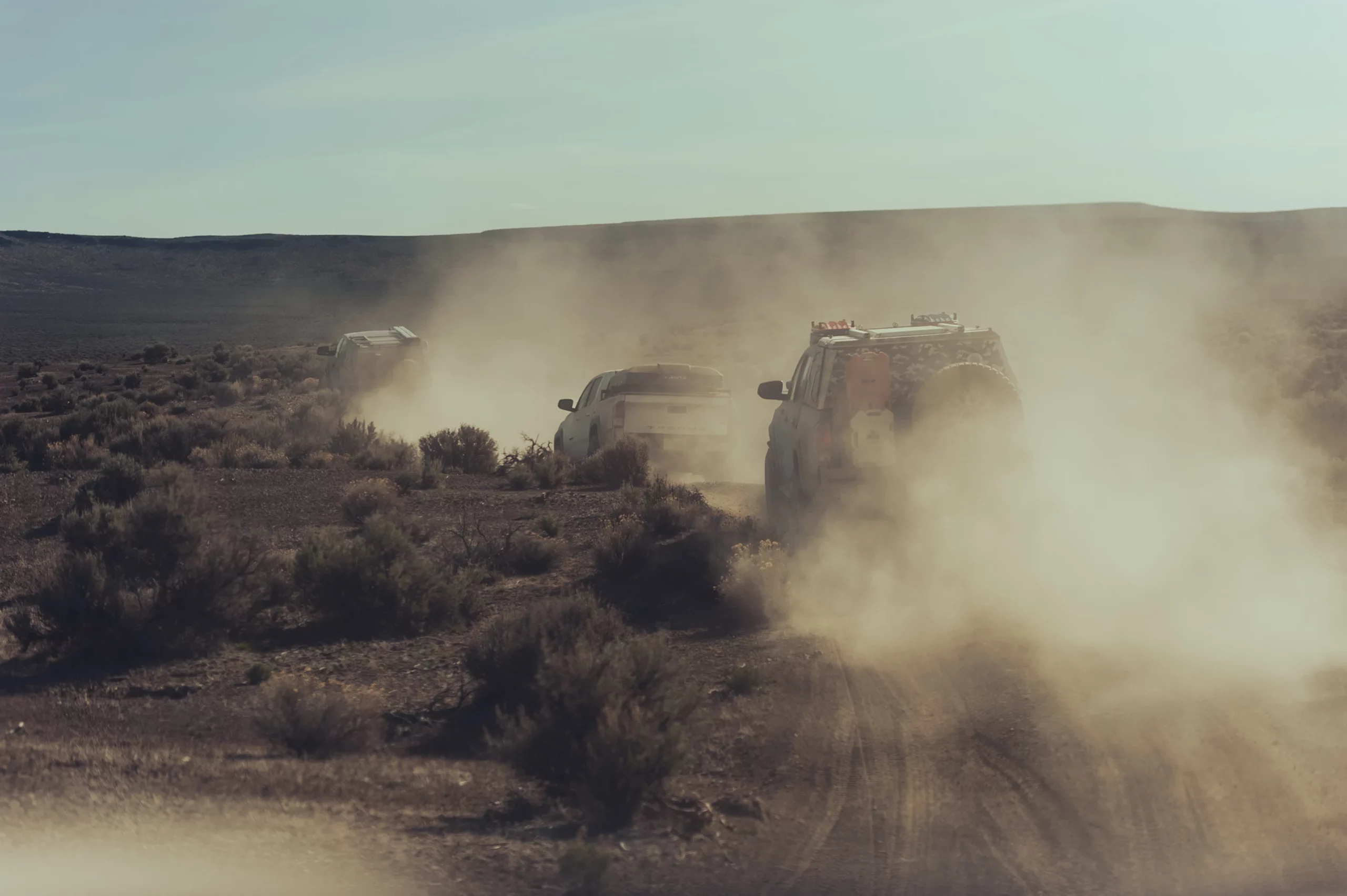 Three off-road vehicles, resembling rugged overland campers, drive through a dusty desert terrain, surrounded by sparse shrubs on a sunny day. Dust clouds billow behind the vehicles as they navigate the rugged landscape, creating a sense of movement and adventure.