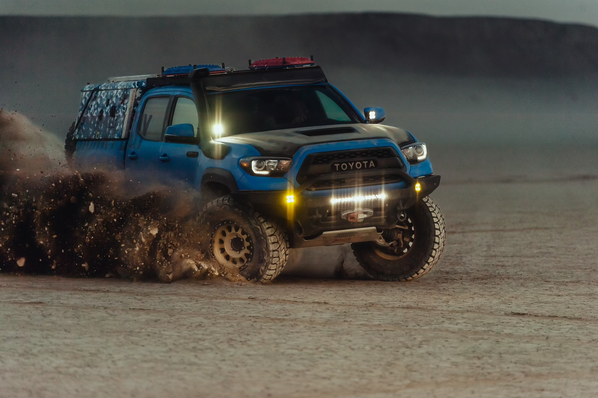 A blue Toyota Tacoma truck with off-road modifications drives through a sandy desert, kicking up dust. Equipped for overland adventures, the truck features roof lights, fender flares, and prominent off-road tires. The background consists of an expansive, barren landscape under an overcast sky.