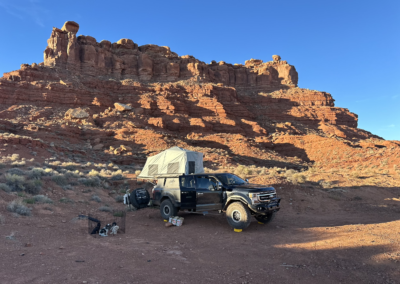 A black overland vehicle with a roof tent, marketed by Skinny Guy, is parked in a desert landscape near a large red rock formation. Camping gear is scattered on the ground, including a camp chair, tools, and a grill. The sky is clear with no visible clouds—perfect for adventurous campers.