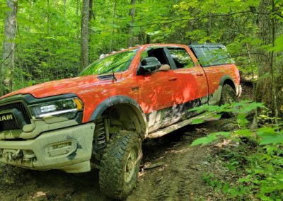 A red RAM off-road truck with mud-covered tires and sides is navigating through a dense, green forest on a muddy trail. The truck's front wheels are elevated on a rough, uneven terrain as it maneuvers like an overland champ. Sunlight filters through the leaves, illuminating the scene.