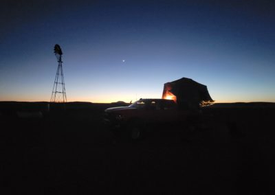 A silhouette of a pickup truck with a rooftop tent illuminated from within set against a twilight sky. The horizon has a warm glow, and there's a windmill visible on the left side of the scene where overland campers gather. A single star is visible in the dusky sky.