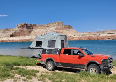 A bright orange pickup truck with a rooftop tent is parked on grassy terrain beside a serene blue lake. Barren, scenic rock formations rise in the background under a clear blue sky. Overland campers would envy its setup, perfect for an adventurous Skinny Guy ready to explore the wild.