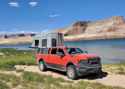 A red Ram pickup truck with a Skinny Guy elevated tent on the back is parked near a picturesque lake surrounded by rocky cliffs and hills. The scene is under clear blue skies with sparse clouds. The sandy and grassy shoreline adds to the rugged overland setting.
