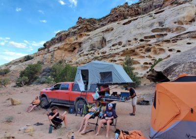 A group of overland campers enjoys a camping trip in a rocky desert landscape. A red truck with a Skinny Guy rooftop tent setup is parked next to an orange ground tent. Campers sit on chairs and prepare food from the truck, surrounded by trees and rock formations under a blue sky.