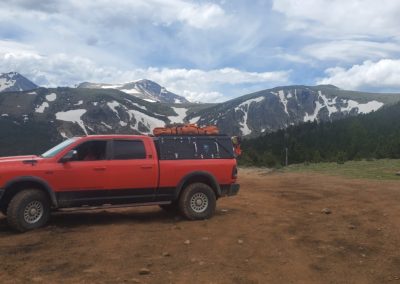A red pickup truck with gear on its roof is parked on a dirt road, overlooking a scenic mountain range with patches of snow and green trees. The sky above is partly cloudy, perfect for overland campers like the Skinny Guy adventurers.