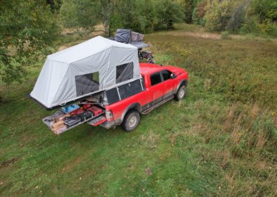 A bright red pickup truck with a tent setup on its bed, perfect for overland campers, is parked in a grassy field surrounded by lush greenery. Camping gear is visible in the truck bed. The scene is set in a remote, nature-filled location.