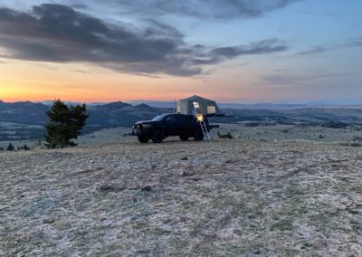 A black overland truck with a rooftop tent is parked on a hillside at sunset. The tent is illuminated, indicating someone might be inside. The landscape features rolling hills and mountains in the distance under a partly cloudy sky. A single tree stands nearby, perfect for Skinny Guy campers exploring the terrain.