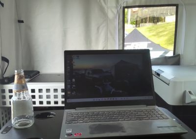 A camper inside a tent-like structure is using a laptop placed on a black surface labeled "Lagun." There is a bottle of sparkling water next to the laptop, and a window in the background shows an overland scene with grass and trees.