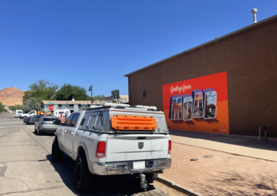 A white truck with equipment on top is parked on a street. In the background, a mural on a brown wall reads "Greetings from Moab" with scenic illustrations in each letter. The scene is set under a clear blue sky.