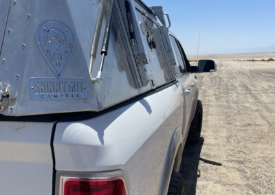A white truck with an aluminum Skinny Guy Campers unit on the back is parked on a barren, sandy landscape under a clear blue sky. The camper has a logo and appears rugged, suggesting it's designed for off-road adventure.