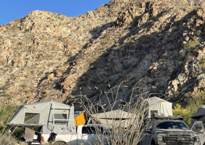 Rugged desert landscape with rocky hills under a clear blue sky. A campsite with two vehicles is in the foreground, both equipped with rooftop tents. Sparse vegetation and cacti are scattered around the site.