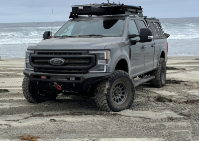 A gray Ford F-Series Super Duty, perfect for overland adventures, is parked on a sandy beach with waves in the background. The truck is equipped with large off-road tires, a roof rack holding various gear, and an attached Skinny Guy camper. The sky is overcast.