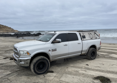 A white pickup truck with a camper shell is parked on a sandy beach. The ocean and rocky shoreline are visible in the background under a cloudy sky.