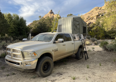 A four-door pickup truck parked in a rugged outdoor setting, equipped with an elevated tent set up on its roof. Skinny Guy campers would love the surrounding landscape of rocky hills, sparse trees, and clear skies, perfect for overland adventures in a remote area.