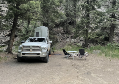 A white pickup truck with a rooftop tent is parked in a wooded area. Adjacent to it, there’s a campsite setup with two folding chairs and a table near a fire pit. Tall trees and rocky terrain surround the site.