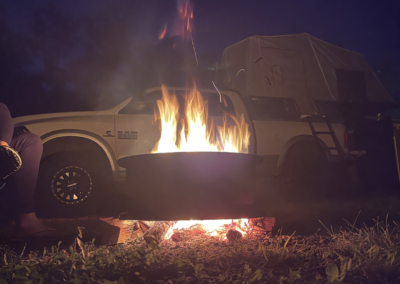 A campfire burning brightly in the foreground with smoke rising. In the background, there's a white pickup truck with a rooftop tent. It's nighttime, and the scene is lit by the glow of the fire.