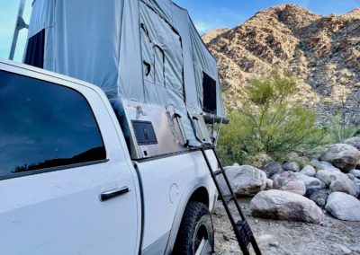 A white pickup truck is parked in a rocky desert area, boasting a rooftop tent mounted on its bed, perfect for overland campers. A ladder provides access to the cozy setup while arid vegetation and a mountainous background stretch under the clear blue sky.