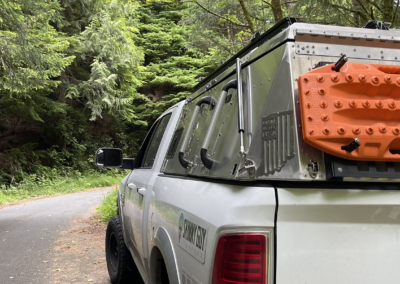 A white pickup truck with an off-road recovery board mounted on the back is parked on a narrow forest road. The truck, ideal for overland campers, is surrounded by dense, lush green trees and vegetation, creating a serene and adventurous atmosphere.