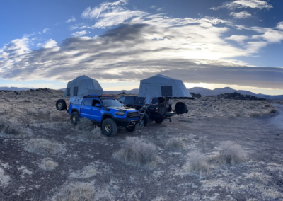 A panoramic view of two overland vehicles with rooftop tents set up in a vast, rocky desert landscape. The sky is partly cloudy with the sun peeking through, casting light on the surrounding terrain and distant mountains. A dirt road extends into the distance on the right, perfect for adventurous campers.