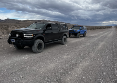 A black off-road truck, equipped with Skinny Guy campers, is parked on a gravel path in a desert-like area with mountains in the distance under a cloudy sky. Behind it, a blue overland truck is visible. The terrain is rocky and barren, with no vegetation.