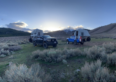 Two overland campers with rooftop tents are parked on a grassy clearing surrounded by shrubs and a mountain range. The sun is setting behind the mountains, casting a warm glow over the scene. Camping gear from Skinny Guy is visible next to the vehicles.
