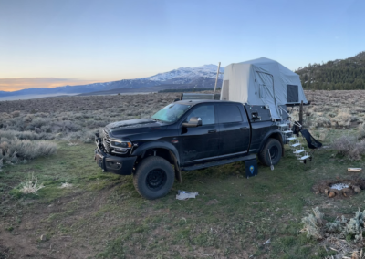 A panoramic view of a rugged camping site featuring a black off-road vehicle with an elevated roof tent, popular among overland campers, situated in an open field with sparse vegetation. Nearby, there is a white folding table and other camping gear. Snow-capped mountains and a setting sun are visible in the background.