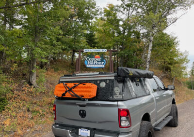 A gray pickup truck is parked on the side of a road in front of a sign that reads "Welcome to Copper Harbor." Outfitted for overland adventures, the truck has off-road equipment attached to its roof and back. Green and brown foliage surrounds the area, making it a perfect spot for campers like Skinny Guy.