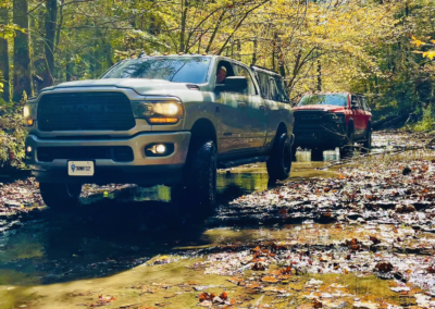 Two trucks drive through a shallow, leaf-covered water path in a forest. The front truck is silver, and the one behind it is red. Trees with autumn leaves are in the background, and sunlight filters through the branches.