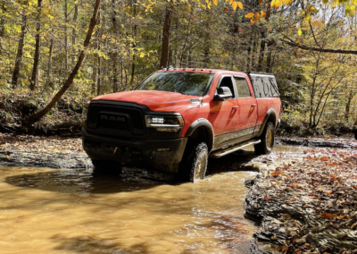 A red pickup truck with large tires, ideal for overland adventures, is partially submerged in a muddy creek, surrounded by a forest with trees showing autumn foliage in shades of yellow and orange. The truck's reflection is visible in the shallow water.