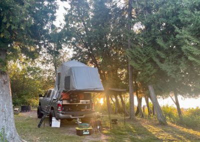 A truck with a roof tent set up in a forested campsite showcases the ideal overland adventure. There's camping gear, including boxes, chairs, and a small cooking area, around the truck. The sun is shining through the trees, casting a warm glow over the scene, inviting adventurous campers to relax and enjoy.