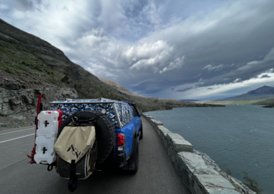 A blue SUV with overlanding gear, including spare tire, fuel, and supplies, parked on a mountain road. It overlooks a vast lake under a dramatic cloudy sky with rugged hills in the distance.