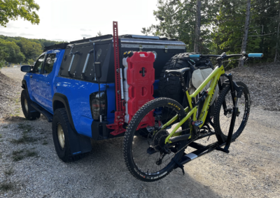 A blue off-road truck parked on a gravel road, equipped with a roof tent and carrying a yellow mountain bike on a rear rack. Red fuel containers and spare tires are attached to the back. Trees and greenery are visible in the background.