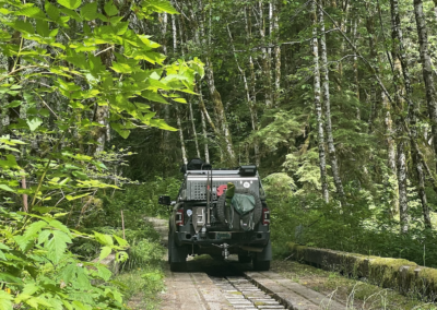A rugged, overland vehicle equipped with gear is driving down a narrow, overgrown dirt path through a dense forest. The vehicle is carrying backpacks and other equipment on the back, suggesting outdoor or adventure travel for seasoned campers. Various green plants and trees surround the path.