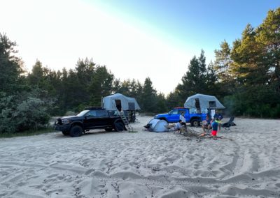 A campsite on a sandy area features two overland vehicles parked, each equipped with rooftop tents. A ground tent sits between them, and three campers are sitting around a campfire while one person stands nearby. Trees frame the background against a clear sky.