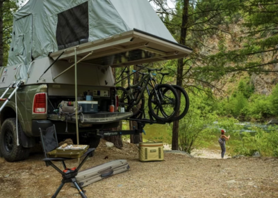 A camping setup with a truck parked in a lush, forested area by a river. The overland vehicle has an elevated roof tent, bicycles mounted on the back, and a variety of camping gear including a chair, cooler, and table. A skinny guy is fishing by the river in the distance.