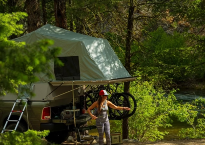 A person stands near a Skinny Guy truck outfitted with a rooftop tent and camping gear in a forested area. A bicycle is mounted on the back of the overland vehicle. The individual appears to be setting up or taking down equipment amidst the lush greenery.