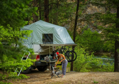 A person wearing a red cap and striped pants is setting up camping gear beside an overland vehicle with a rooftop tent. Bicycles are mounted at the back of the vehicle. The campsite is surrounded by lush green trees with a river flowing nearby, creating an idyllic setting for these adventurous campers.