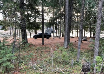 A black camping truck with a rooftop tent is set up in a forest area near a lake. Amidst the campers, the scene is surrounded by tall trees with filtered sunlight shining through. The ground is covered with pine needles and scattered branches, perfect for an overland adventure.