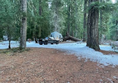 A black truck is parked on a snow-dusted campsite in a dense forest. Behind the truck, a large tent is set up near tall trees. The ground has patches of brown earth and scattered snow, with various camping items around the site, creating an ideal scene for overland campers to enjoy nature.