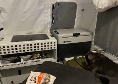 A compact camping setup for overland campers features a portable Dometic refrigerator, shelves with various items, and a white foldable crate. A dog rests on a green mat beside the refrigerator inside a tent with white fabric and attached storage pockets from Skinny Guy.