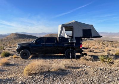 A black pickup truck with an elevated, rooftop tent is parked in an arid, rocky landscape with distant hills and a clear blue sky. A ladder descends from the tent to the ground, showcasing a perfect overland camping setup. Dry vegetation is scattered around the area, ideal for adventurers and campers alike.