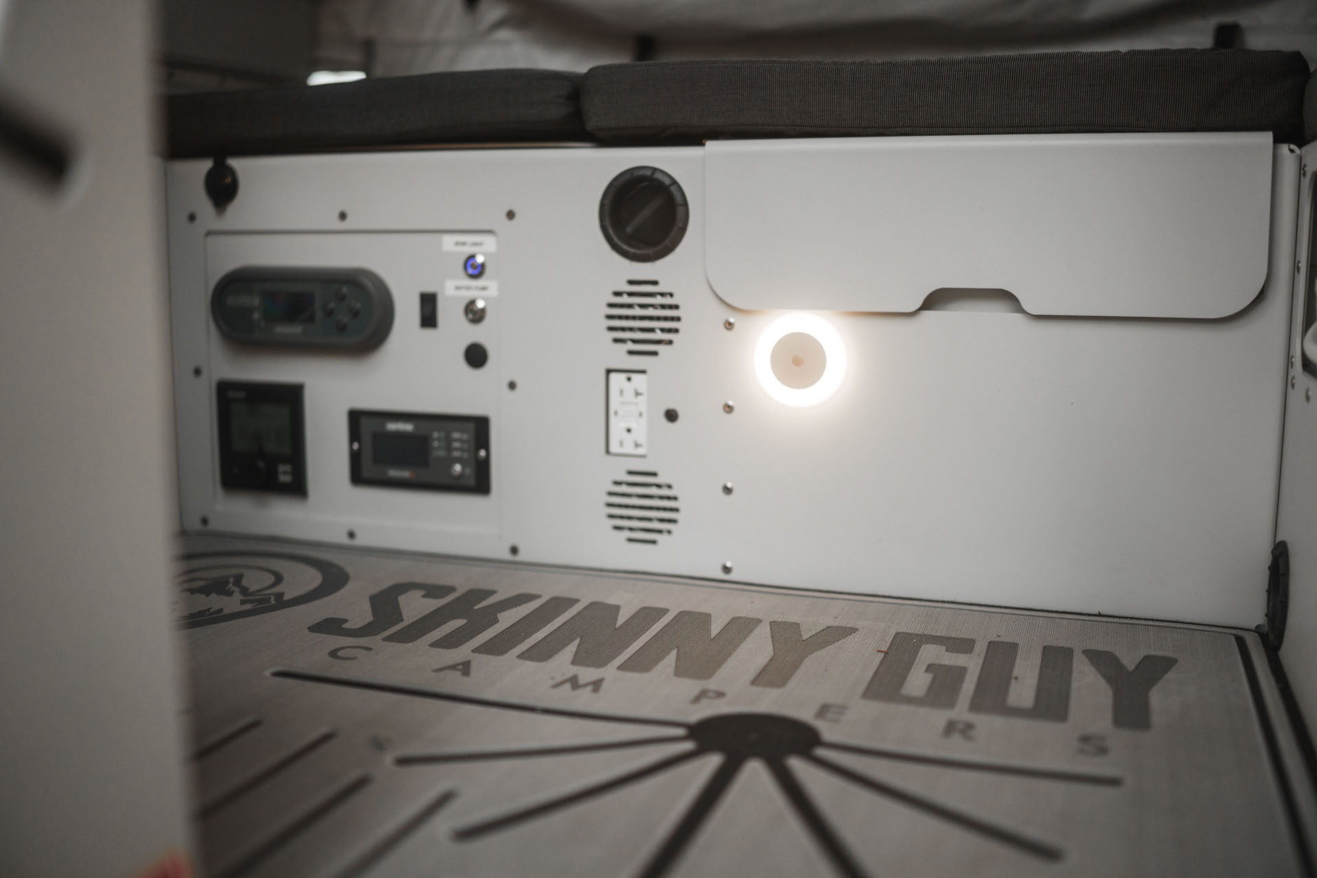 Interior view of a compact, minimalist camper trailer by Skinny Guy Campers. The floor features the "Skinny Guy Campers" logo. There are control panels, switches, and a small light illuminating the wall. A cushioned bench seat is against the back wall, perfect for overland adventures.