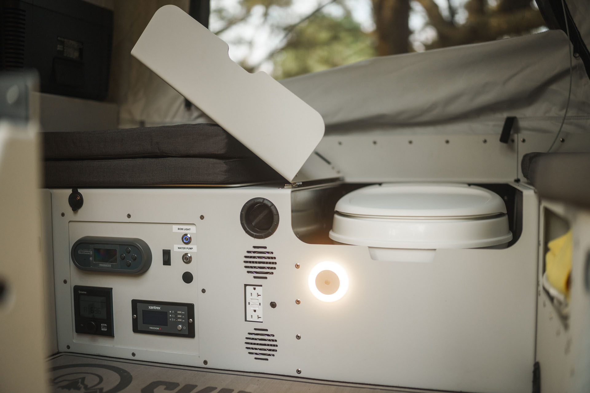 Interior view of a camper van showing a built-in bench seat and a concealed toilet, partially revealed with its lid open. The cushioned bench seat is complemented by various controls and electrical outlets on the side panel, illuminated by a small light—perfect for overland adventures with campers like Skinny Guy.