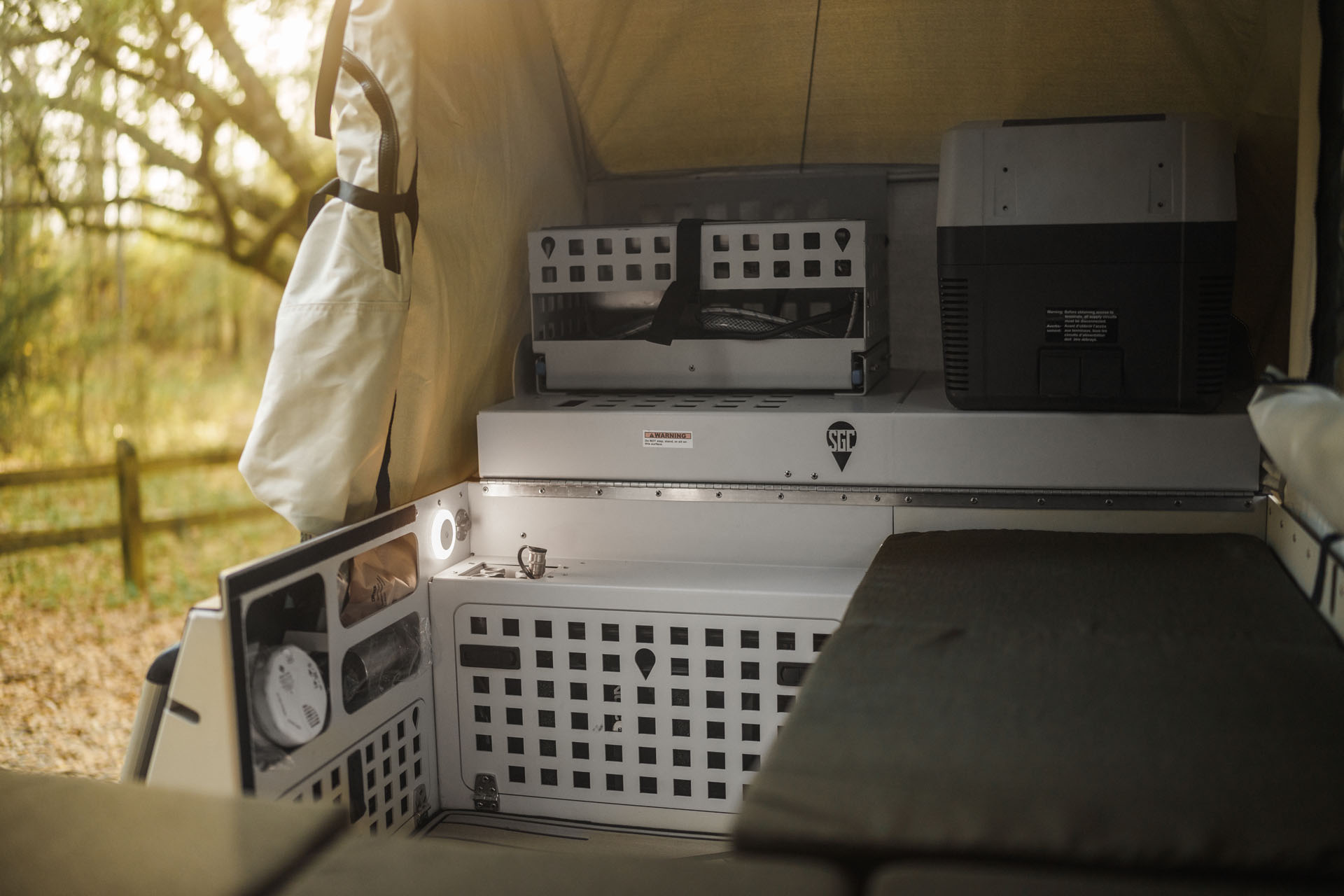 This image showcases the interior of a modern camper van by Skinny Guy, featuring modular storage compartments and a small refrigerator. The setup includes a countertop and drawers for organization. Light streams through an open tent flap, highlighting a rustic overland setting with trees and a fence.