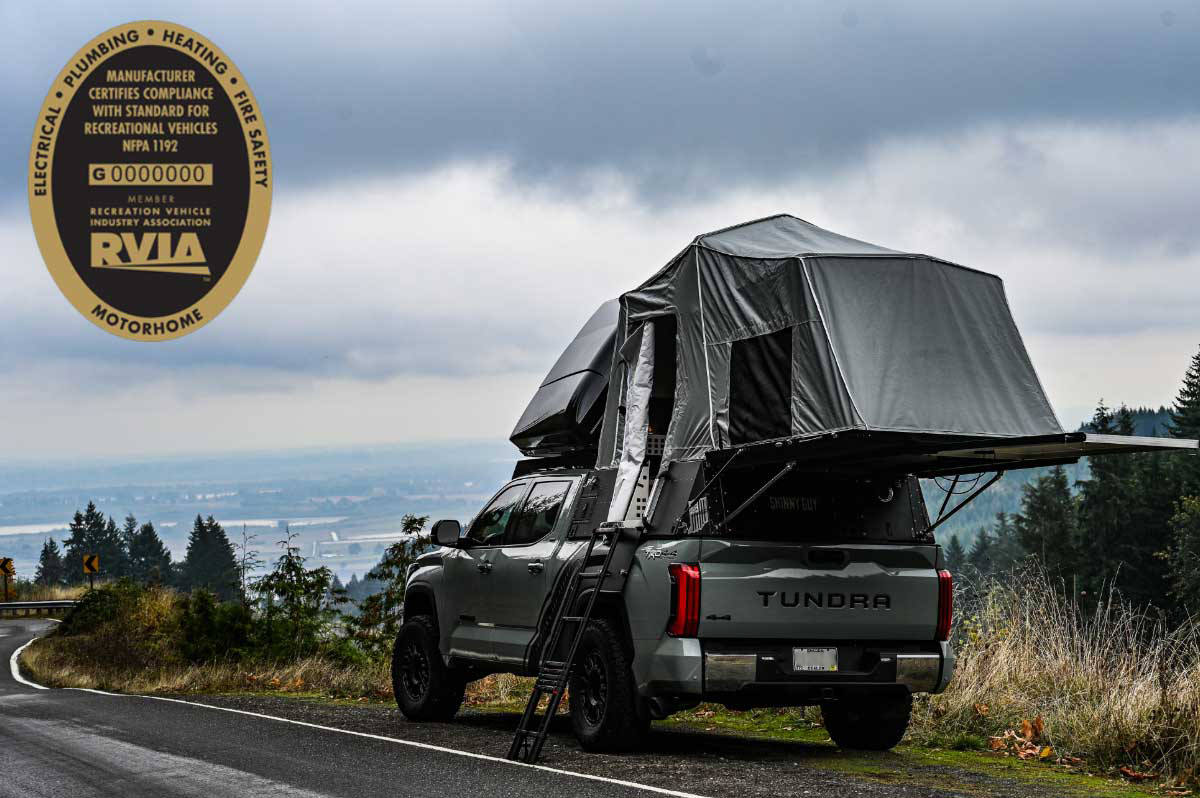 A gray pickup truck with a rooftop tent, perfect for overland campers, is parked on the side of a winding mountain road. The Skinny Guy tent is expanded and ready for use, with a ladder attached. In the background, there's a scenic view of a valley under a cloudy sky. A round certification logo is visible in the top left.