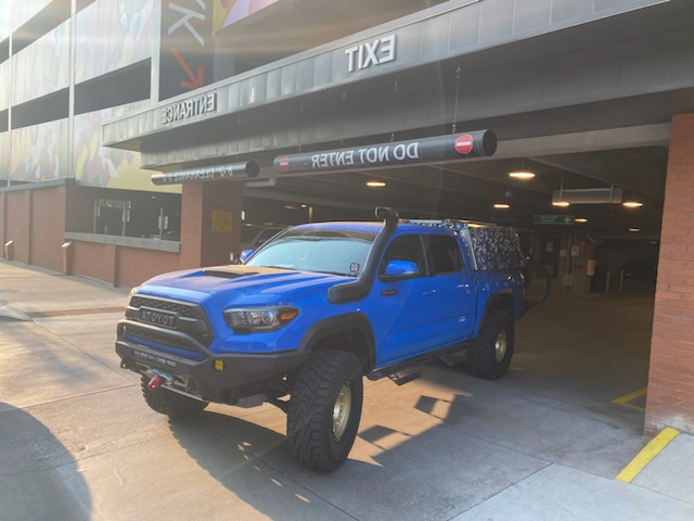 A blue Toyota Tacoma truck with off-road modifications is exiting a parking structure. The truck, ideal for overland adventures, boasts large tires, a front winch, snorkel, and roof rack with a storage box. Skinny Guy campers would envy this setup. The parking structure has 