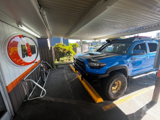 A blue pickup truck, perfect for overland adventures, is parked under an awning next to a white and orange wall with a round sign. Beside it, a bicycle is chained to a rack near the wall. The location appears to be an outdoor area with some greenery visible in the background.
