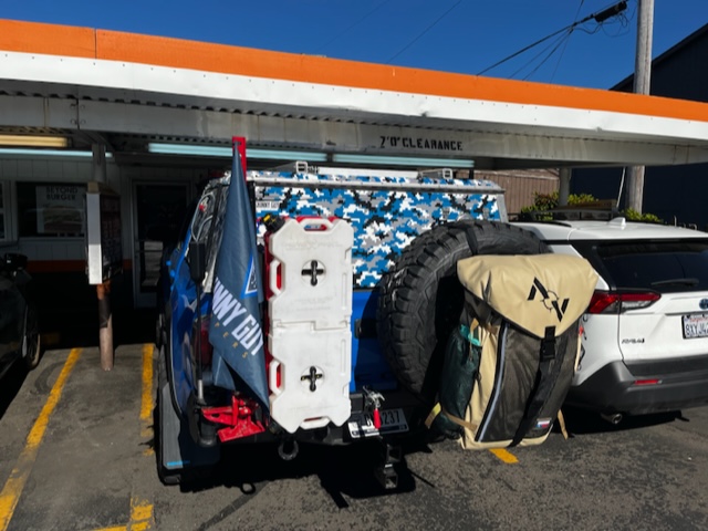 A modified overland vehicle with a blue camouflage design is parked in a lot. It features a large spare tire and storage containers attached to the rear. A flag with text is also affixed to the vehicle. In the background, a building displays a 7'0