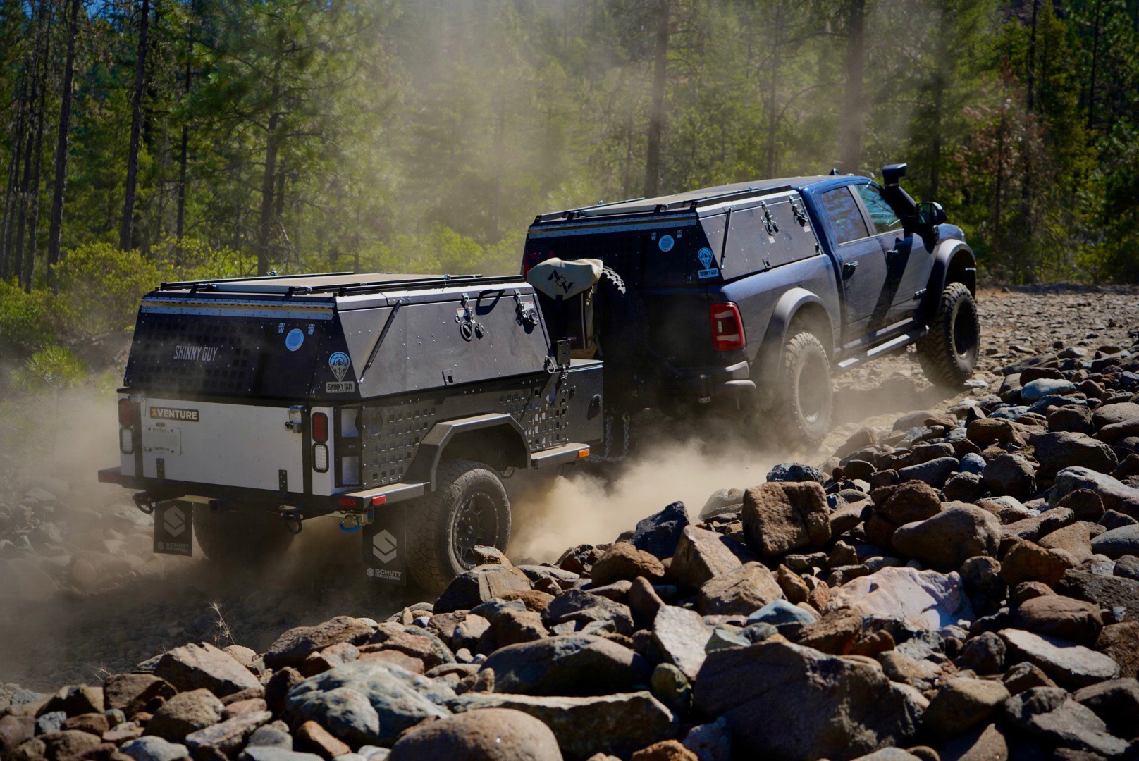 A rugged off-road vehicle towing a Skinny Guy camper trailer navigates a rocky terrain. The scene is set amidst a forest with dust kicked up by the tires, highlighting the challenging and adventurous nature of overland exploration. The vehicle and trailer are equipped for outdoor adventure.